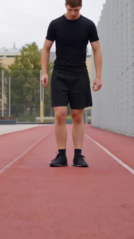 Young male athlete in black sportswear ready to sprint, focuses energy and muscles on the red running track of an urban city stadium during solo fitness session in daylight, slow motion vertical shot.