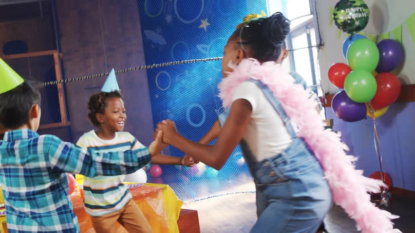 Girl reaching for hands and starting circle, five kids spinning around party table confetti overlay. Children, friends, balloons, hats, celebration, laughter, decor