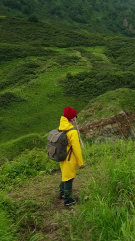 A Hiker in a Vibrant Yellow Raincoat and Red Beanie Gazing at a Majestic Coastal Landscape Surrounded by Lush Greenery and Rugged Cliffs