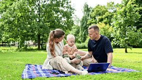 Family couple with little son sitting in park on grass and watching video on laptop. Happy family with child on a stroller outdoors. Mom dad and son relaxing in nature. - Powered by Shutterstock - Get 15% off with code: PIKWIZARD15