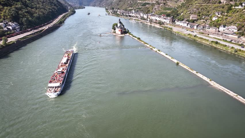 Aerial pull-back shot from Pfalzgrafenstein Castle on its small island in middle of the Rhine River with passing river cruise ship, steep valley slopes, vineyards and riverside town Kaub.