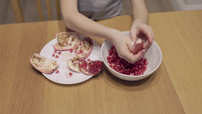 Child carefully removes pomegranate seeds into a plate. Wide top view shows small hands working with bright red fruit and focusing on coordination.
