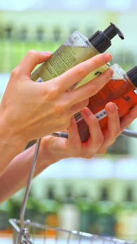 Vertical video: Woman comparing cosmetic product in a store aisle, carefully reading labels and ingredients for the best haircare or skincare choice.