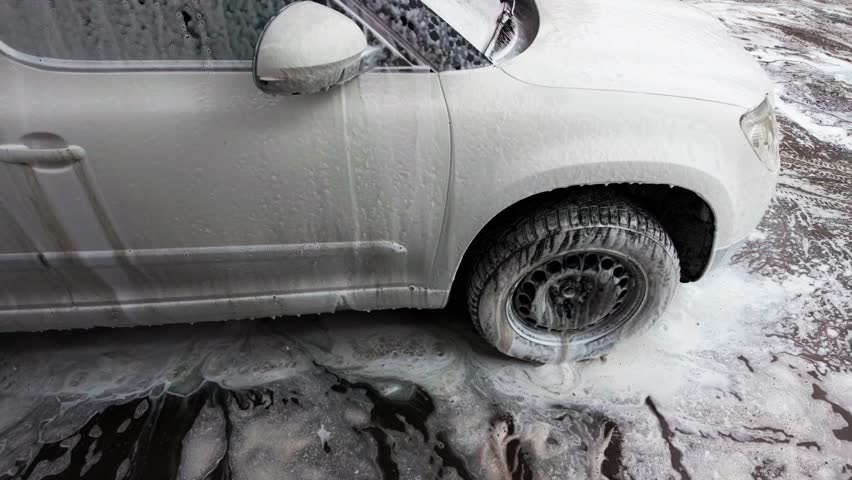 front side suv wheel foam scrub in muddy bay; outdoor detailer working on offroad vehicle, thick suds around fender and rim, splash and runoff across textured driveway, energetic maintenance scene