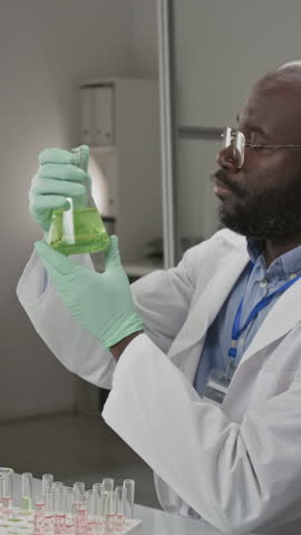 Vertical portrait shot of African American researcher in white coat and protective gloves scrutinizing flask filled with solution in laboratory