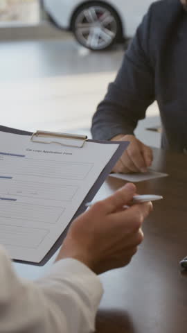 Vertical shot of unrecognizable car dealer giving document to man sitting at desk and signing agreement while buying new car
