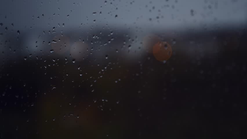 A single raindrop trails down a rain-soaked window during an autumn downpour, creating a soft, moody pattern on the glass. 4K with selective focus.