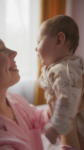 Young cheerful mother smiling, lifting baby up in air, playing with her, rubbing noses and kissing child during joyful moment at home. Vertical clip