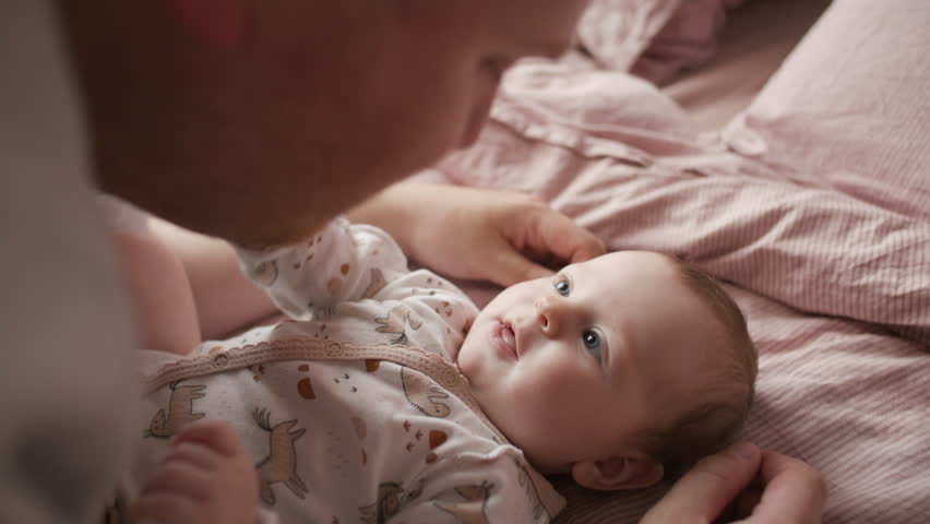 Adorable baby in onesie lying on bed, gazing at loving father leaning above her while bonding at home