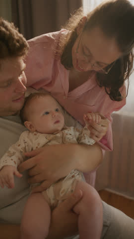 Young happy parents holding their cute baby and sharing kiss while enjoying family time together in cozy living room filled with sunlight. Vertical format footage