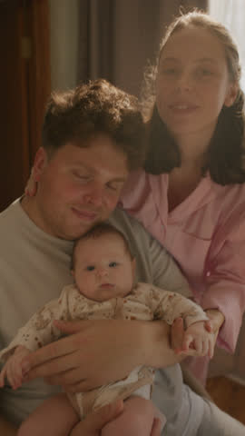 Happy young family holding baby, smiling and looking together at camera in cozy home bathed in warm sunlight. Vertical clip, video portrait