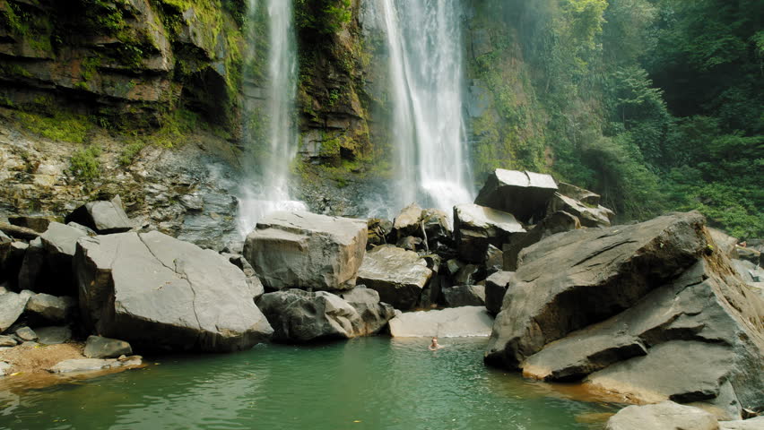Person swimming under refreshing tropical waterfall in natural pool