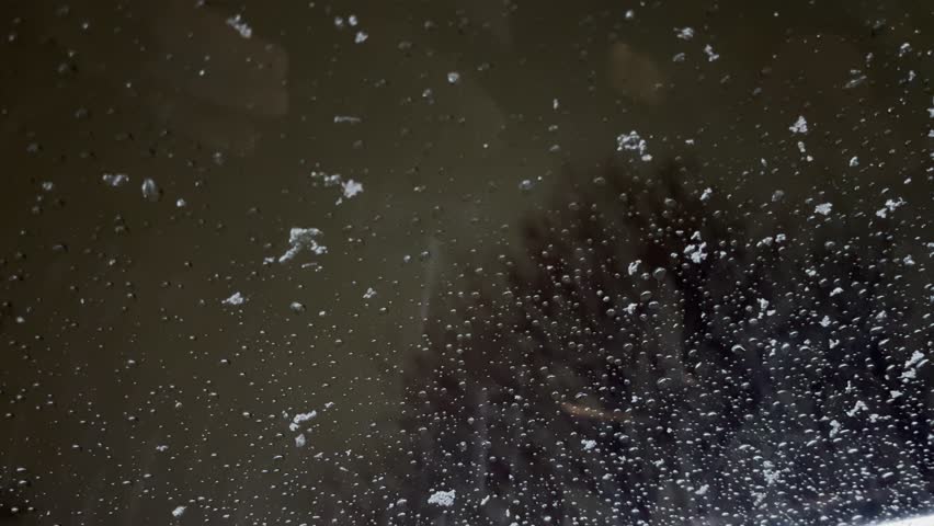 Macro shot of melting snow and sleet accumulating on a glass window at night with blurred warm lights and tree reflection in the dark background.