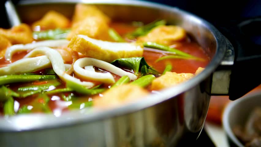 A close up shot shows a simmering metal pot of red Yen Ta Fo, a Thai hot pot filled with squid, fish cake, and water spinach.