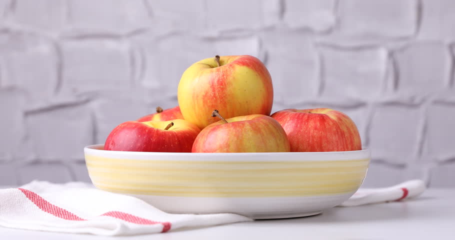 Woman taking ripe red apple from bowl at white table near grey textured wall, closeup