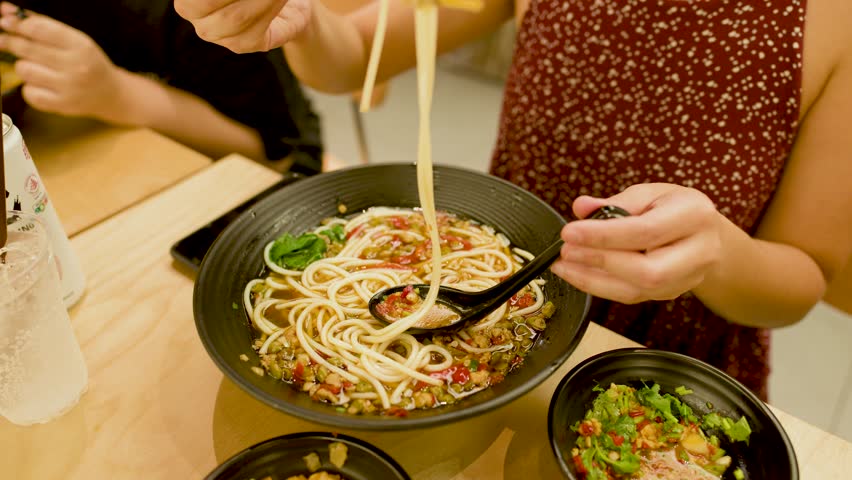 Woman uses chopsticks to lift spicy noodle soup in bright, casual dining setting, overhead view