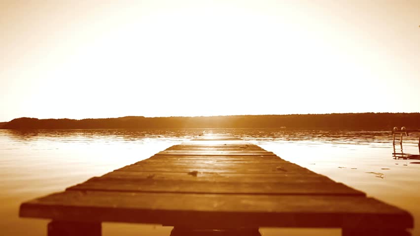 Wooden dock stretching out onto a calm lake at sunset with a golden color tone