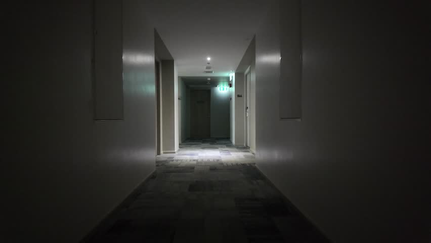 A point-of-view shot of a dimly lit hotel corridor with gray walls and patterned carpet leading towards an exit sign.