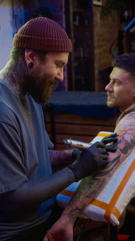 Vertical medium shot of friendly young Caucasian male tattooist chatting with male client while drawing tattoo sleeve on his arm