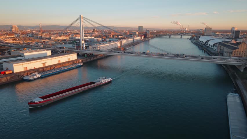 Golden Hour Aerial of Mannheim Rhein Bridge: Barge Navigating Industrial Harbor and City Skyline Traffic at Sunset, Germany