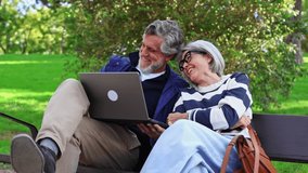Cheerful elderly couple sitting together on a park bench. The mature man and woman are smiling and laughing while using a laptop computer - Powered by Shutterstock - Get 15% off with code: PIKWIZARD15