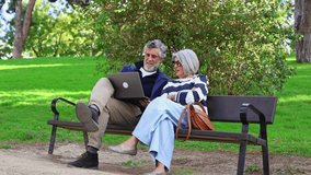 Stylish mature couple sitting on a park bench, happily talking and using a laptop together. Modern seniors enjoying their leisure time outdoors - Powered by Shutterstock - Get 15% off with code: PIKWIZARD15