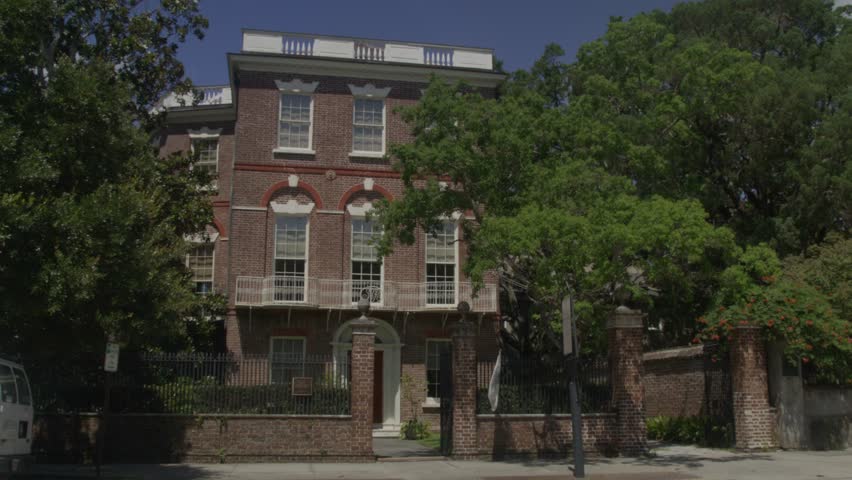 Daytime shot of a historic brick home in Charleston, SC, framed by trees and an ornate iron gate on a sunny summer day with stable video.