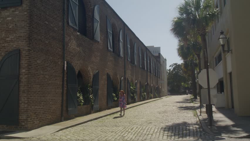 A woman walks along a sunny cobblestone street in downtown Charleston, SC, passing historic brick buildings and palm trees on a bright summer day with stable video shot.