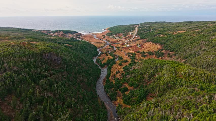 Aerial view of a valley where a winding river passes farms, houses and a road toward the ocean; forested hills and green fields under a bright sky.