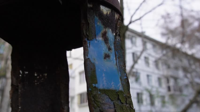 An old rusty metal bird feeder hangs from a tree and sways in the wind. Blue side part is visible on the feeder. Old houses and branches of dry trees are visible behind the feeder.High quality footage