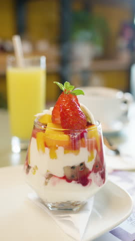 Close-up of a yogurt parfait with fresh fruit and syrup, accompanied by a glass of juice and a cup of hot coffee in the background.