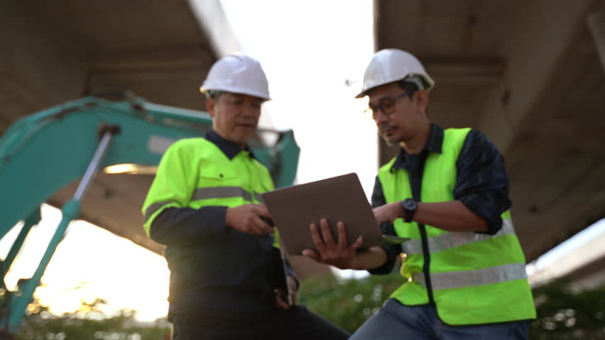 Two construction workers engaged in a discussion while using a laptop under a bridge, highlighting teamwork and technology in urban construction projects.
