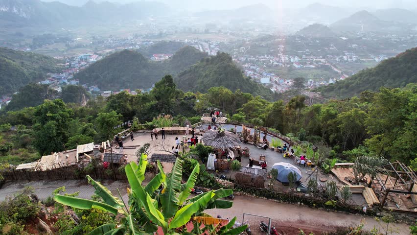 Drone orbit shot around an outdoor hilltop café in Mu Nau, Moc Chau, overlooking a panoramic view of the town below with houses scattered among rolling hills.