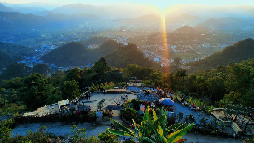 Drone forward shot flying from behind the hilltop café in Mu Nau, Moc Chau, toward the town below, capturing a warm backlit scene with sunlight flaring into the camera.