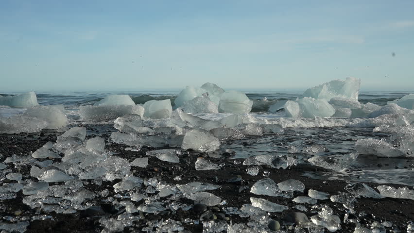 Slow-motion capture of arctic sea waves crashing onto glacier ice fragments on Diamond Beach, Iceland. Desolate black sand contrasts with nature's raw, spectacular show of ice and ocean.