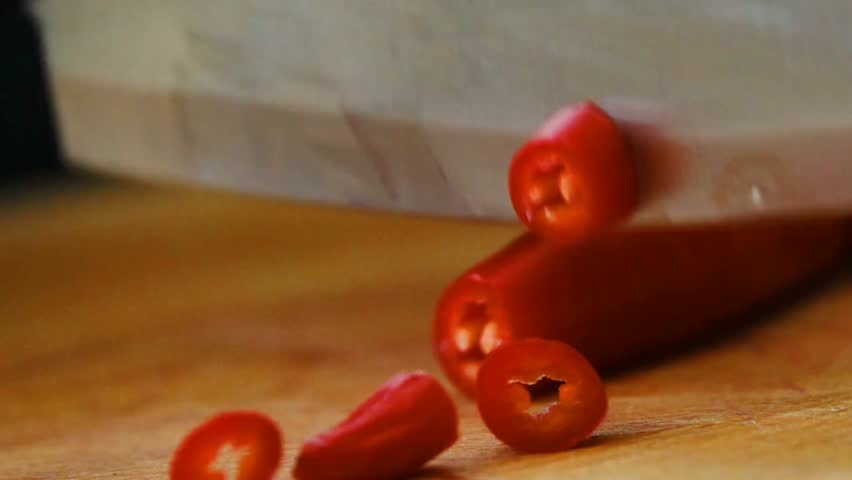 Close-up of a knife slicing a red chili pepper on a wooden board, highlighting cooking, food prep, and culinary action.