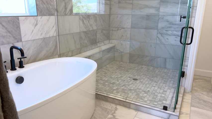 Wide shot of a bathroom with full marble tile walls and windows. Scene includes a freestanding tub and modern matte black fixtures.