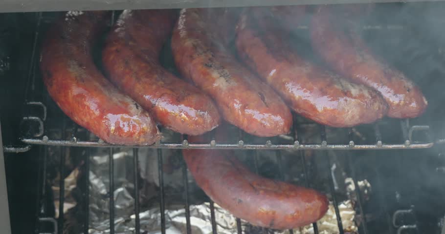 A closeup shot of a juicy smoked sausages (Bratwurst sp.) cooking on metal rack inside barbecue smoker filled with smoke