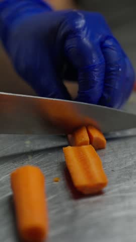 A gloved hand slicing a carrot into pieces on a metal counter while preparing fresh vegetables