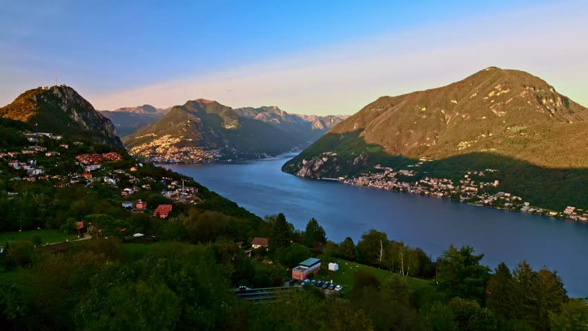 Carona, Switzerland – September 25, 2023: Panoramic view over Lake Lugano toward Monte Brè and Monte Caprino, with hillside homes and distant alpine peaks under warm evening light.
