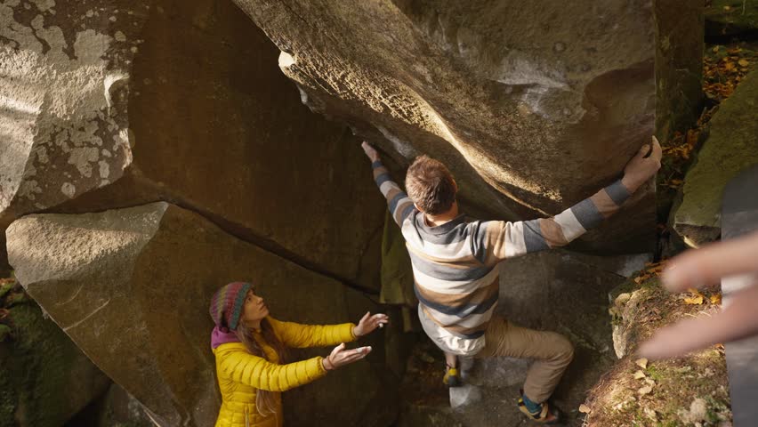 man rock climber climbs on overhanging boulder by it edges using wide hands grips, friends belay him. climbing outdoor in autumn forest on natural rocks. healthy active lifestyle, training outdoors.