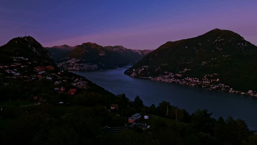 Carona, Switzerland – September 25, 2023: Lake Lugano at dusk with Monte Brè and Monte Caprino silhouetted against a fading violet sky near the Italian border.