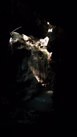 Fast-flowing water cascades over and between large, wet boulders in a dark, rugged gorge opening, with sunlight dappling the rocks.