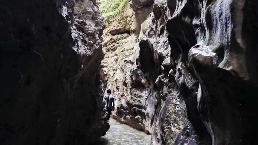 A narrow, dark cave passage opening to a slice of light, showing rippling water flowing between two imposing, rugged rock walls.