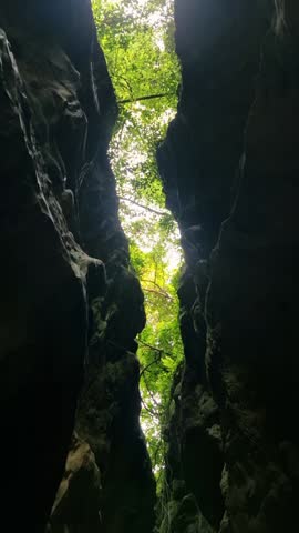A narrow, dark cave passage opening to a slice of light, showing rippling water flowing between two imposing, rugged rock walls.