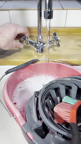 Vertical video - Closeup of a man’s hand holding onto a flowing kitchen sink tap or faucet, of steaming hot water, into a car wash bucket of soapy suds water.
