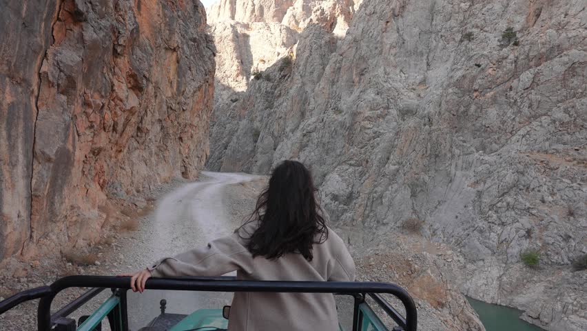 A woman rides in a safari through Kemaliye Canyon, lifting her arms while enjoying dramatic rocky cliffs, a river landscape and a bridge in the background, capturing a lively and scenic adventure travel moment.