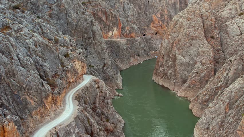 A dramatic view of the river flowing between steep rocky walls in Kemaliye Canyon. A narrow cliffside road curves along the canyon, with a small riverside platform visible below the rugged terrain.