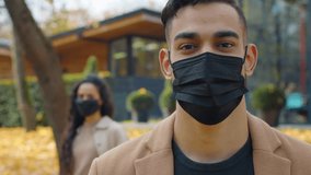 Hispanic man in medical face mask looking at camera posing in foreground in city autumn park unrecognizable woman in background Arabian male guy portrait keep distance prevention coronavirus covid19 - Powered by Shutterstock - Get 15% off with code: PIKWIZARD15