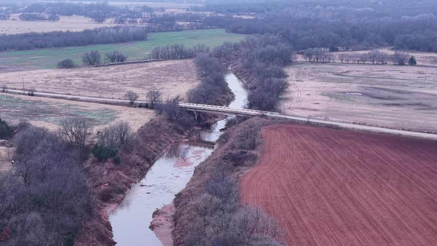 Aerial drone view of a winding river, country bridge, red soil farmland, and winter fields in rural Oklahoma with bare trees and soft overcast light. Scenic quiet landscape.